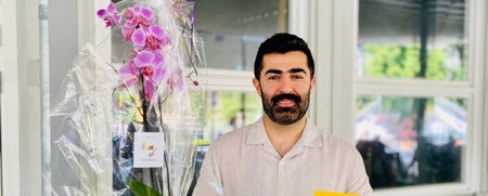 Man med mörkt hår och skägg håller en blomma i kruka och ett gult kort med texten 'TACK!'.