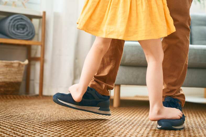 Ett barn med gul kjol och bara ben dansar med en vuxen genom att stå på en vuxens fötter. De dansar på en brun matta.