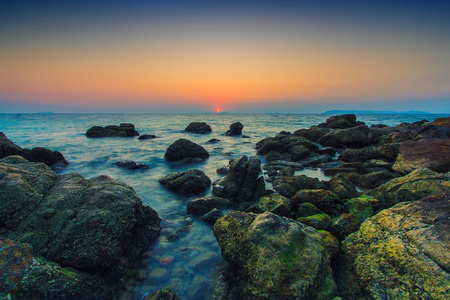 Solnedgång över ett stenigt hav med klippor i förgrunden och färgglad himmel.