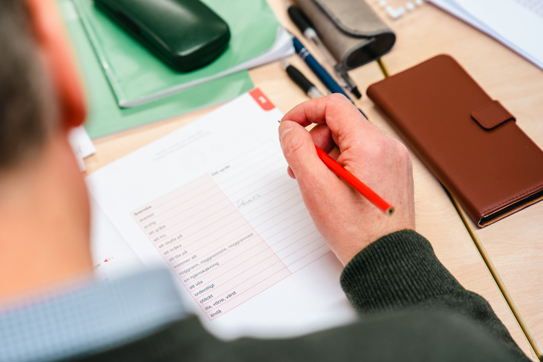 En person skriver med röd penna på ett papper vid ett skrivbord med anteckningsblock, pennor och fodral.