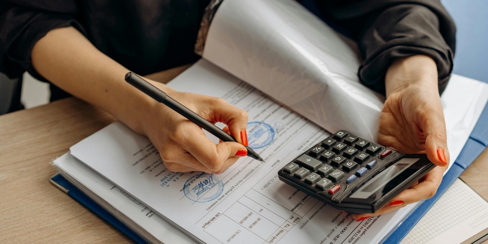 En person skriver på dokument med en penna och håller en miniräknare vid ett skrivbord.
