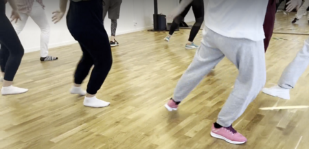 En grupp människor dansar i en ljus dansstudio med trägolv. Fokus på ben och fötter.