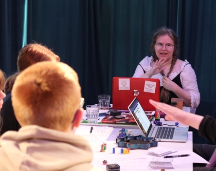 En grupp personer spelar rollspel runt ett bord med tärningar, papper och en laptop. En kvinna leder spelet.
