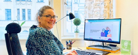 Kvinna med glasögon sitter vid skrivbord och dator i ljust kontorsrum, ler mot kameran.