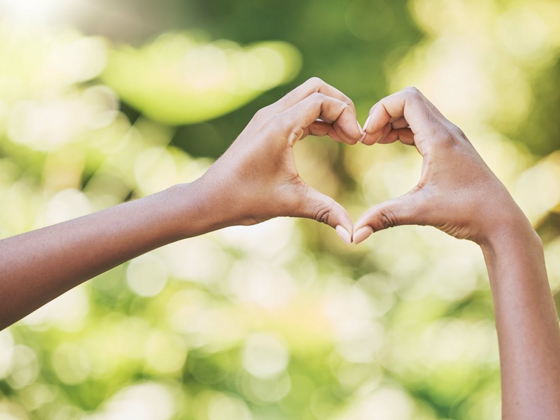 Två händer formar ett hjärta mot en suddig, grön bakgrund utomhus.