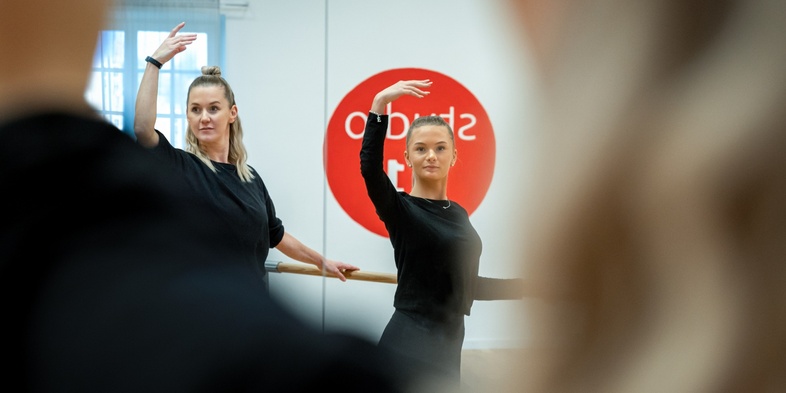 Två kvinnor övar dans framför en spegel i en dansstudio, båda med en arm upplyft.