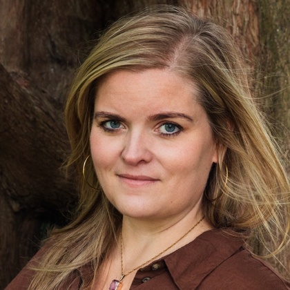 Person med blont hår och brun skjorta står framför ett stort träd. 