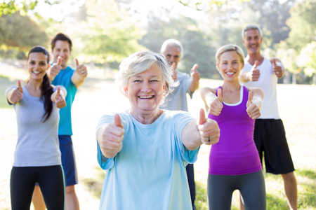 En grupp glada människor utomhus ger tummen upp mot kameran, med en äldre kvinna i fokus längst fram.