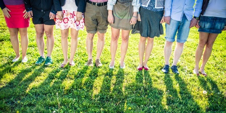 Åtta personer står på gräs, endast deras ben och fötter syns, med långa skuggor i solskenet.