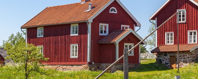 Två röda trähus med vita knutar på landet, omgivna av gräs, staket och blå himmel.