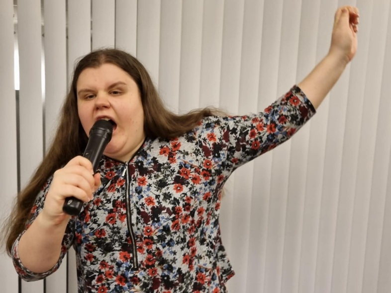 Kvinna med långt hår sjunger i mikrofon och poserar framför vita persienner.