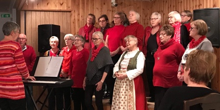 En kör står på en liten scen och sjunger, ledd av en dirigent vid ett elpiano. Publik sitter framför.