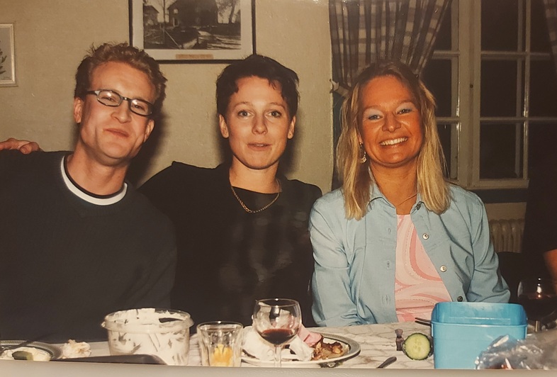 Tre personer sitter vid ett matbord och ler mot kameran under en middag inomhus.