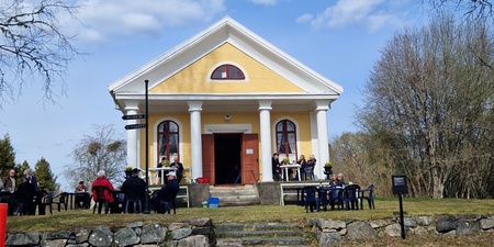 En gul byggnad med vita pelare, omgiven av människor som sitter utomhus på stolar under en klarblå himmel.