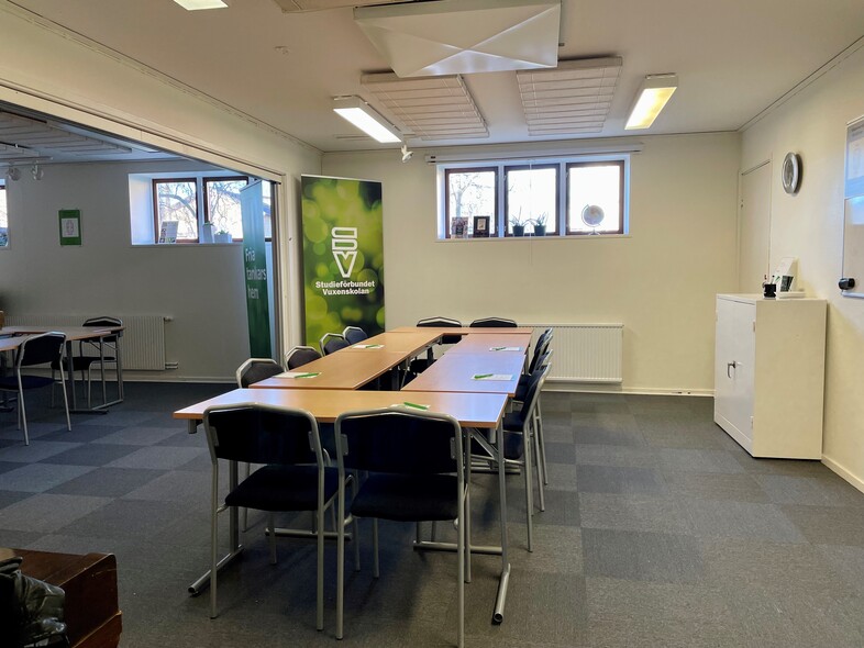 Ett ljust konferensrum med bord och stolar, fönster, en grön skylt och en vit förvaringsmöbel.