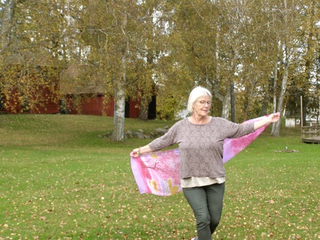 Äldre kvinna med vitt hår går på gräsmatta och håller ut en rosa sjal, höstträd i bakgrunden.