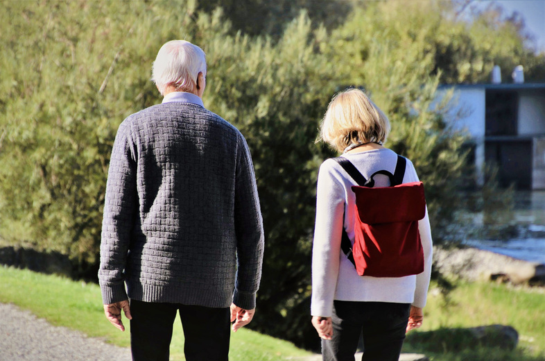 Två äldre personer promenerar utomhus på en stig, sedda bakifrån, med grönska i bakgrunden.