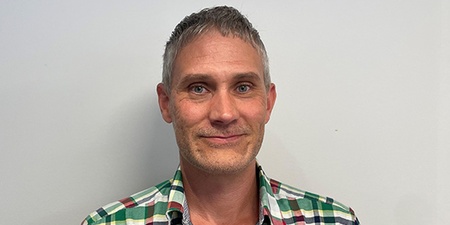 Person med kort hår och rutig skjorta står mot en ljus vägg.