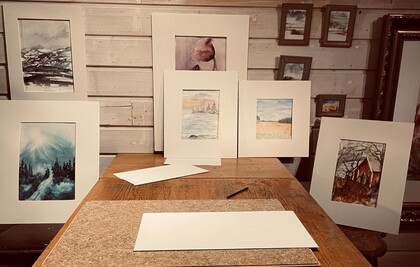 Ett bord med flera inramade akvarellmålningar uppställda mot en trävägg, samt papper och penna på bordet.