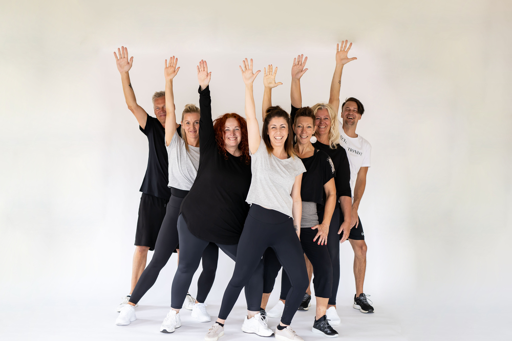 Sju vuxna personer står i rad och poserar glatt med ena armen uppsträckt mot vit bakgrund.