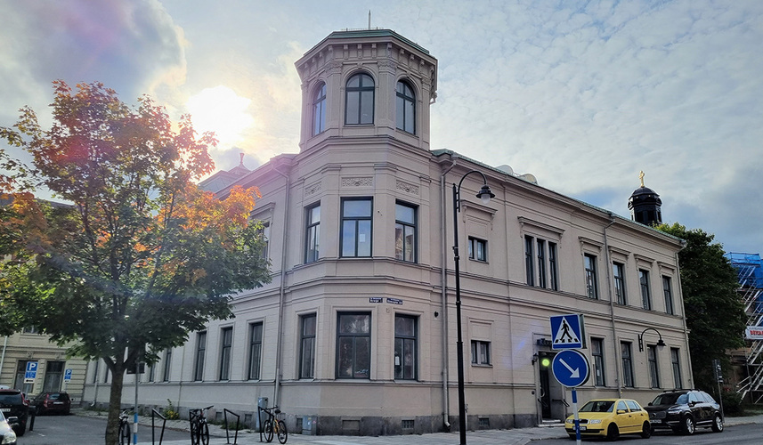 Gammal ljusgrå byggnad med torn, träd och bilar i förgrunden, sol bakom moln.