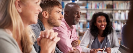 Fyra vuxna personer sitter vid ett bord i ett bibliotek och arbetar tillsammans med datorer och anteckningar.