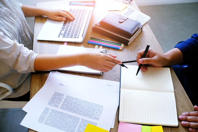 Två personer arbetar tillsammans vid ett skrivbord med laptop, anteckningsblock och dokument.
