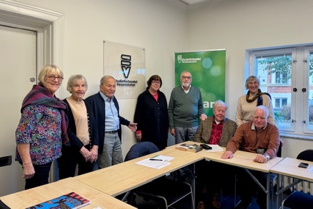 En grupp äldre personer står och sitter runt ett konferensbord i ett mötesrum.