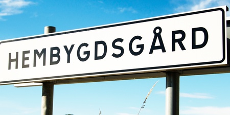 Vägskylt med texten 'Hembygdsgård' mot en lantlig bakgrund med blå himmel och fält.