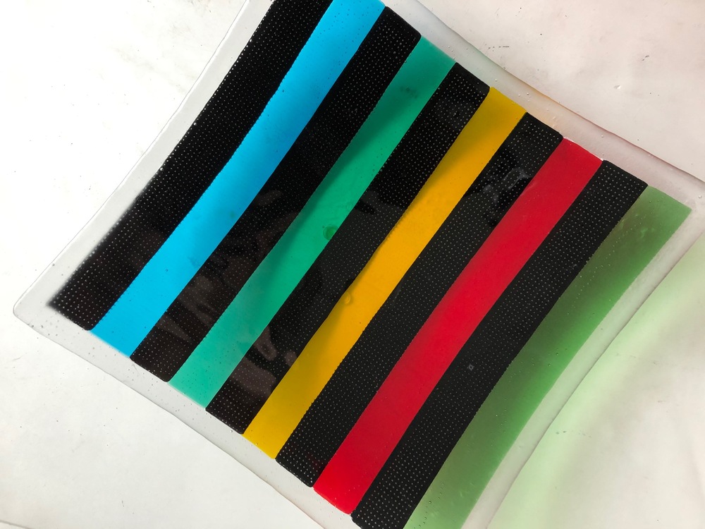 En rektangulär glasskiva med färgade ränder i svart, blått, grönt, gult och rött på vit bakgrund.