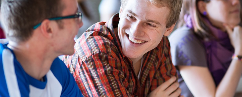 Tre unga vuxna sitter vid ett bord i ett klassrum och samtalar, två av dem ler mot varandra.