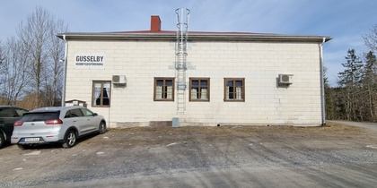 Vit byggnad med skylten 'Gusselby Hembygdsförening', tre bilar parkerade framför, omgiven av träd.