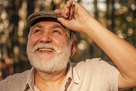 Leende äldre man med vitt skägg och keps står utomhus i soligt skogsparti.