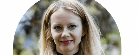 Kvinna med långt blont hår, gul blommig klänning och svart choker, utomhus i grön miljö.