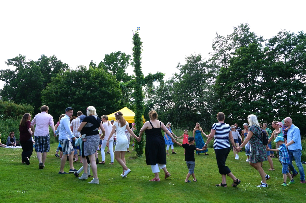 En grupp människor dansar i ring utomhus runt en midsommarstång på en gräsmatta.