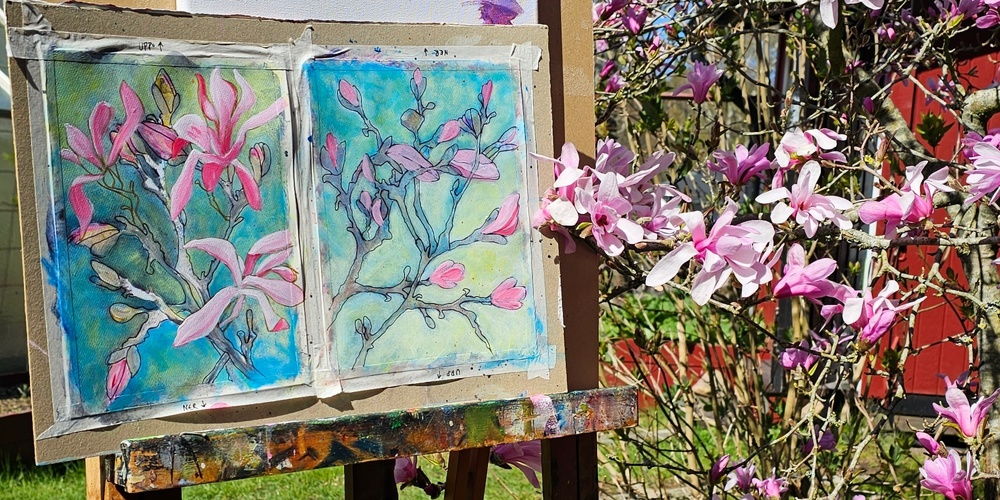 Ett staffli med målningar av rosa magnoliablommor står utomhus bredvid ett blommande magnoliaträd.