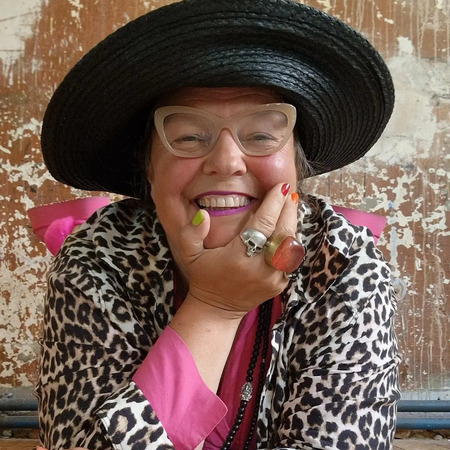 Leende person med stor svart hatt, glasögon och leopardmönstrad jacka mot sliten vägg.