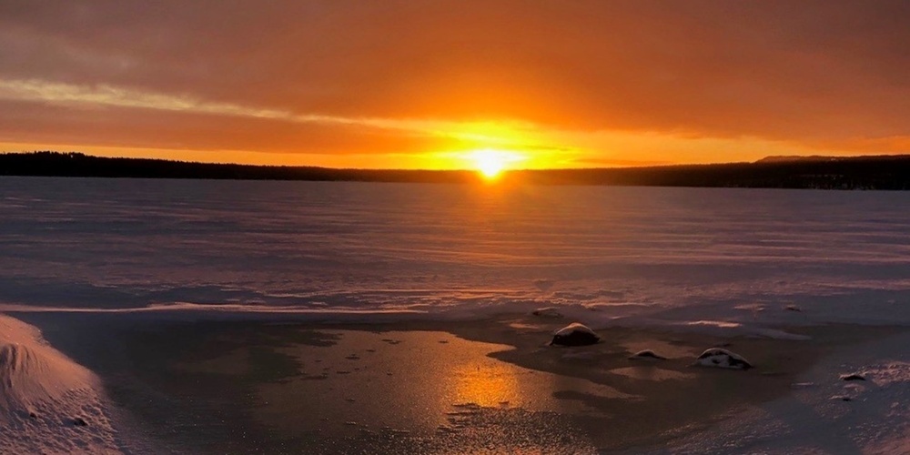 Solnedgång över ett snötäckt landskap med fotspår som leder mot en isvak.