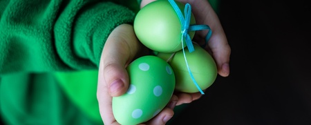 Ett barn i grön tröja håller tre gröna påskägg, ett med prickar och ett med blått band, i sina händer.