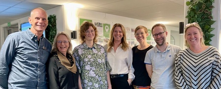 Sju personer står tillsammans och ler i ett ljust kontorslandskap med gröna växter på väggarna.
