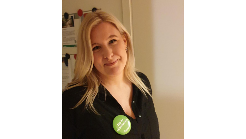 Kvinna med blont hår och svart skjorta står inomhus och ler mot kameran. Hon bär en grön knapp på bröstet.