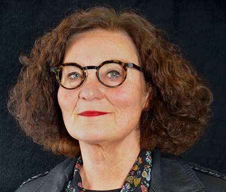 Person med lockigt brunt hår, svart läderjacka och blommig skjorta mot svart bakgrund.