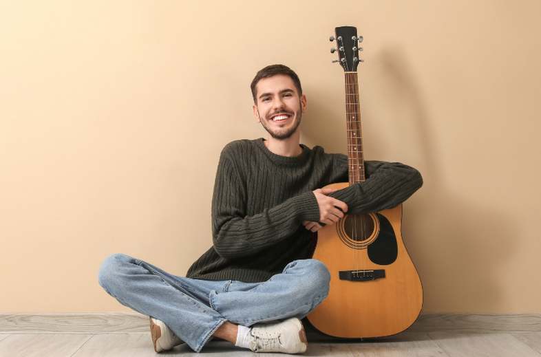 Person sitter på golvet mot en beige vägg med en akustisk gitarr.