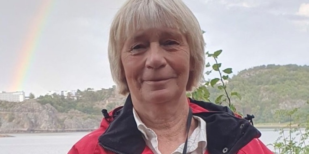 Person i röd jacka med kikare runt halsen står utomhus vid vatten, med regnbåge i bakgrunden.