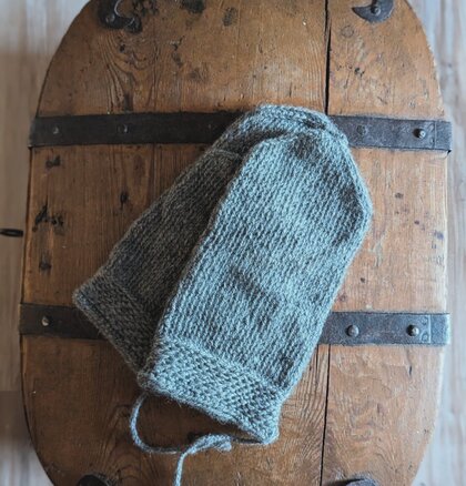 Ett par stickade grå vantar ligger på ett gammalt träbord med metallbeslag.