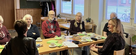 En grupp äldre personer sitter runt ett bord i ett ljust rum och samtalar över fika.