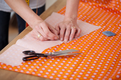 Händer som nålar fast mönsterpapper på orange tyg med sax och krita bredvid.