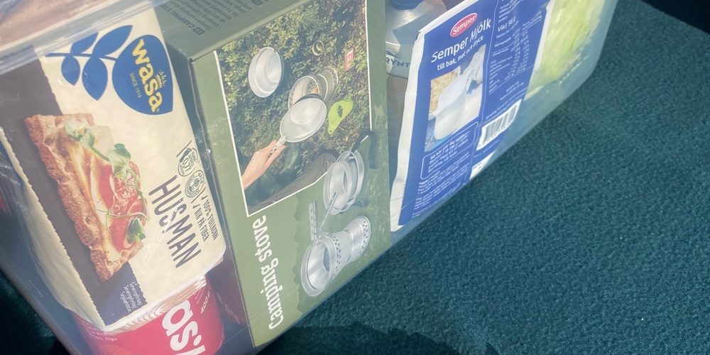 En genomskinlig plastlåda fylld med matvaror och hushållsartiklar står på ett bord utomhus.