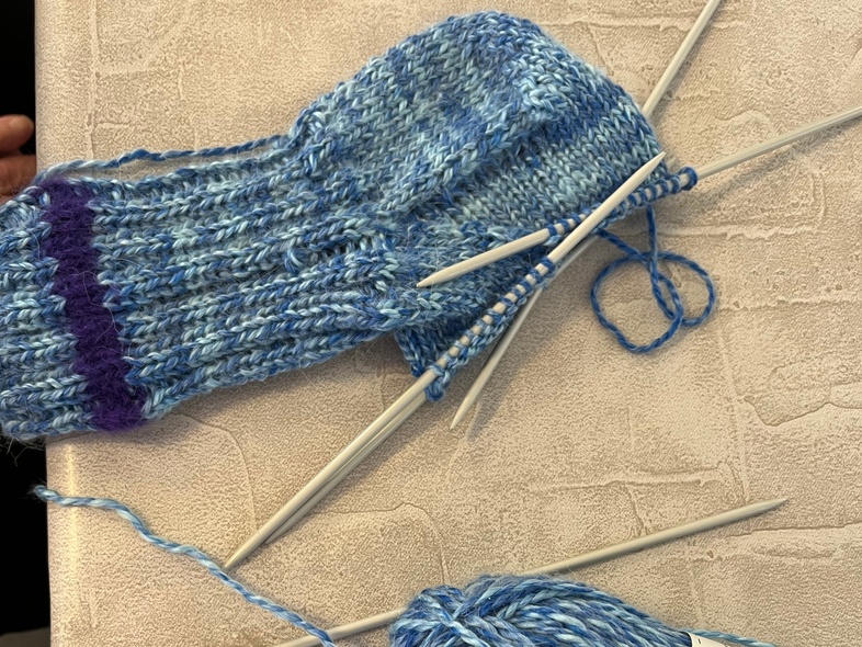 En stickad blå socka under arbete med strumpstickor och ett nystan blått garn på ett bord.