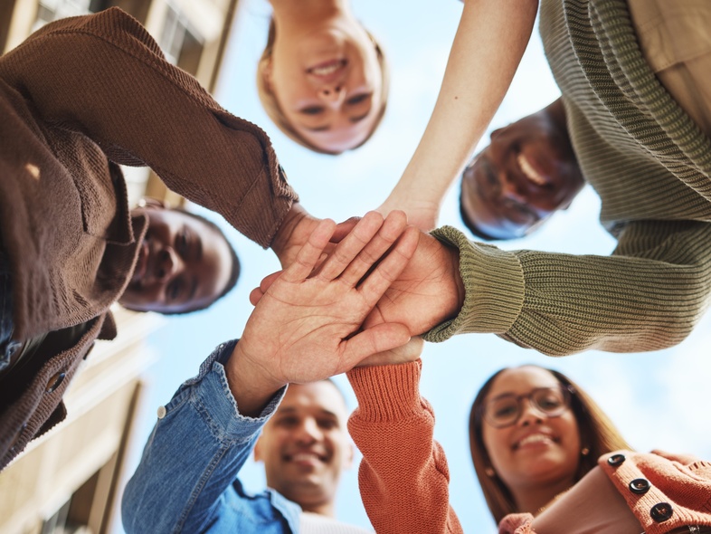 Fem personer står i en cirkel och lägger sina händer ovanpå varandra, sedda underifrån mot himlen.